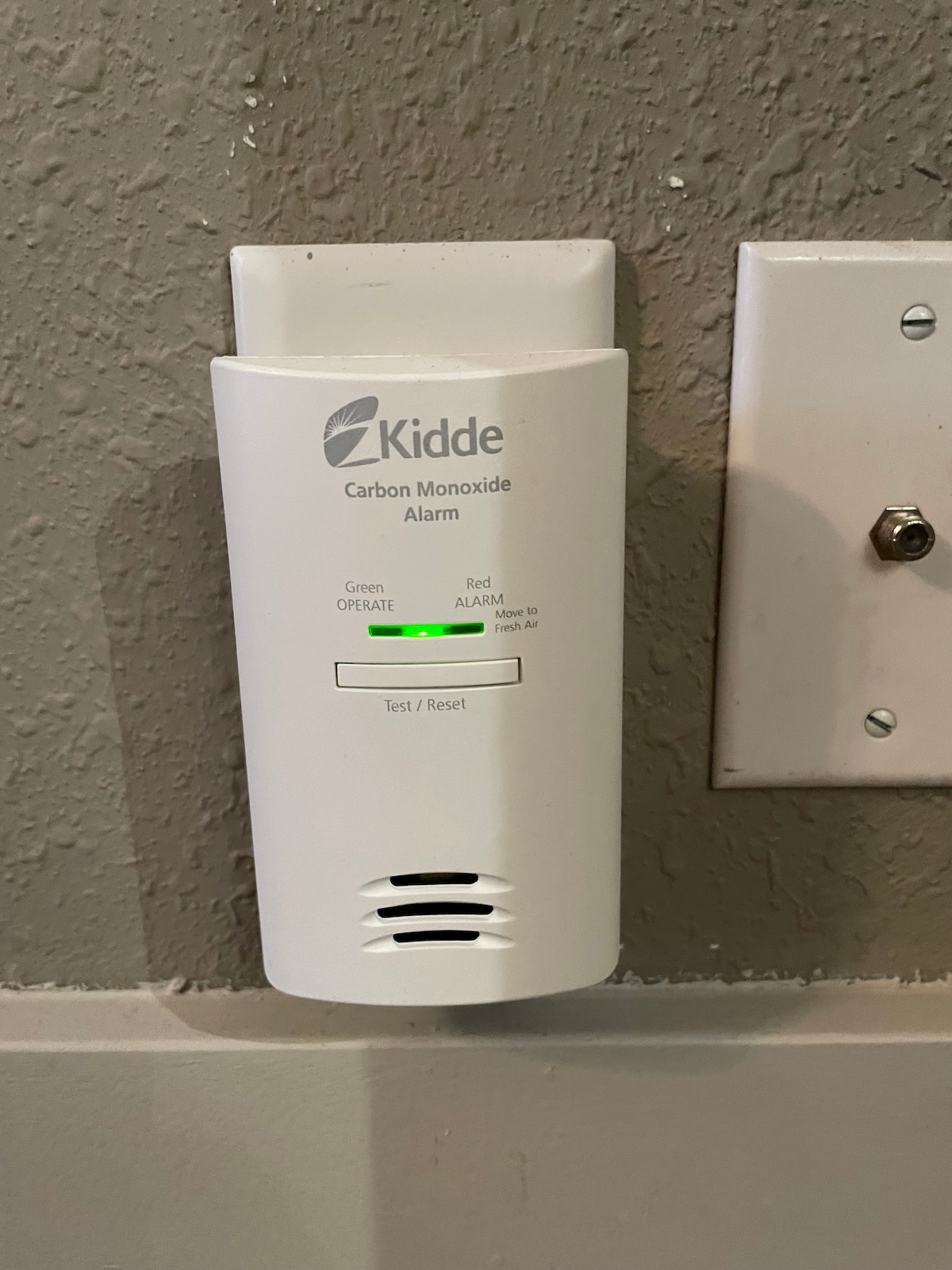 A carbon monoxide alarm is mounted on a textured gray wall. It features a green indicator light showing normal operation. A nearby electrical outlet is visible to the right, with a silver screw for securing the plate. The design emphasizes safety within the rental unit.