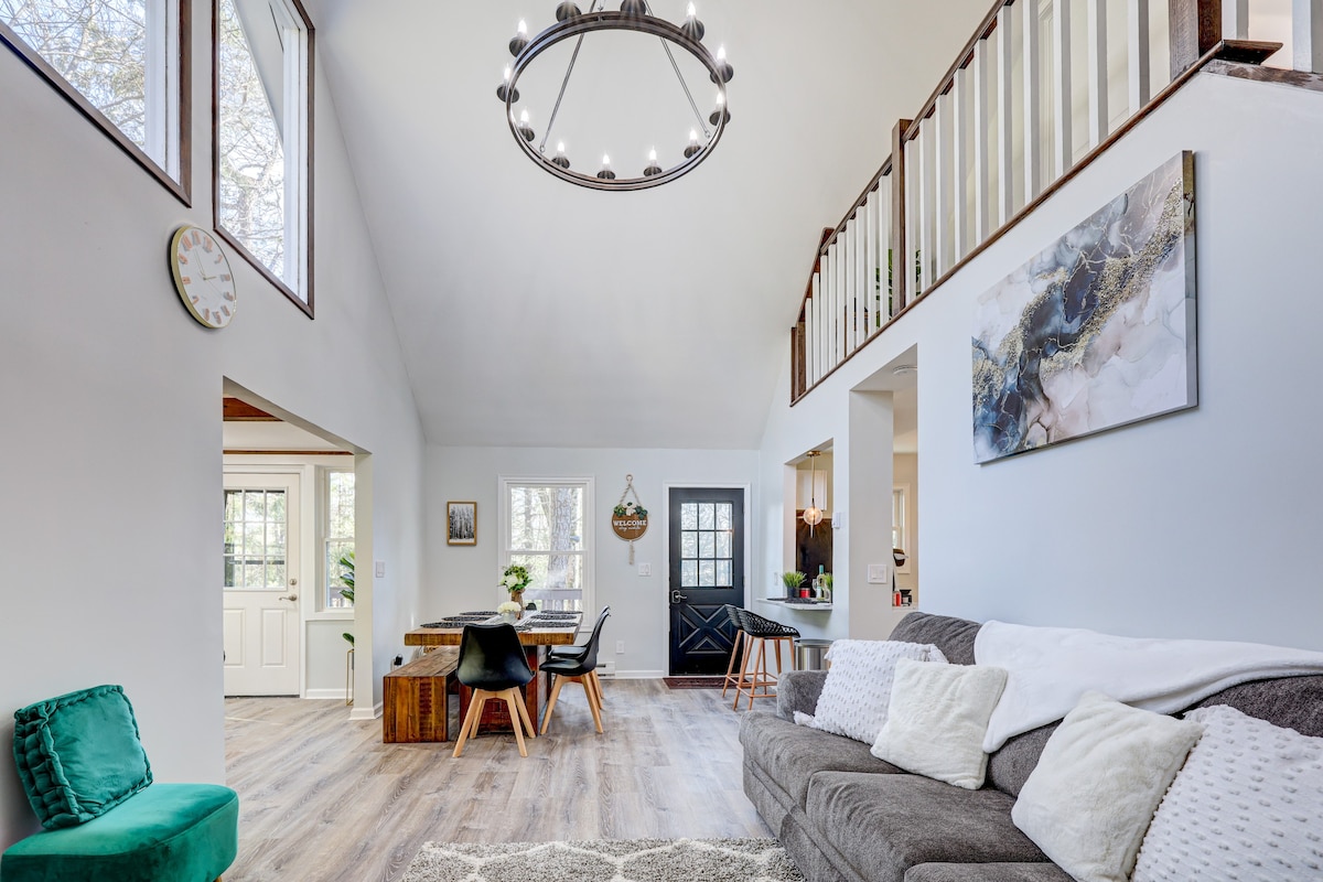 A spacious living area features high cathedral ceilings, complemented by large windows that provide natural light. A comfortable sofa is positioned on one side, while a dining table with black chairs is visible. A modern chandelier hangs above, adding a stylish touch to the room.