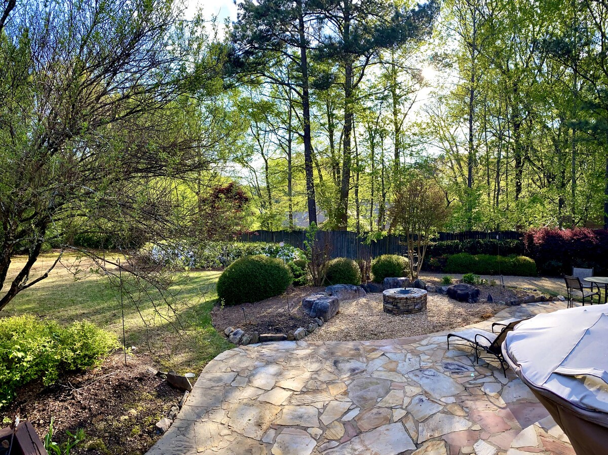 A spacious backyard is revealed, featuring a stone patio that leads to a firepit surrounded by seating. Lush greenery and various shrubs enhance the natural setting, with trees providing dappled sunlight. The area invites relaxation and gathering, offering ample space for outdoor activities.