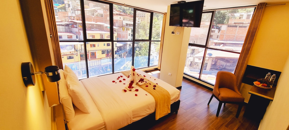 A well-lit room features a spacious bed adorned with decorative petals. Large windows offer views of the surroundings, while a television is mounted on the wall. A chair and small table provide additional seating. Natural light fills the space, enhancing the welcoming atmosphere.