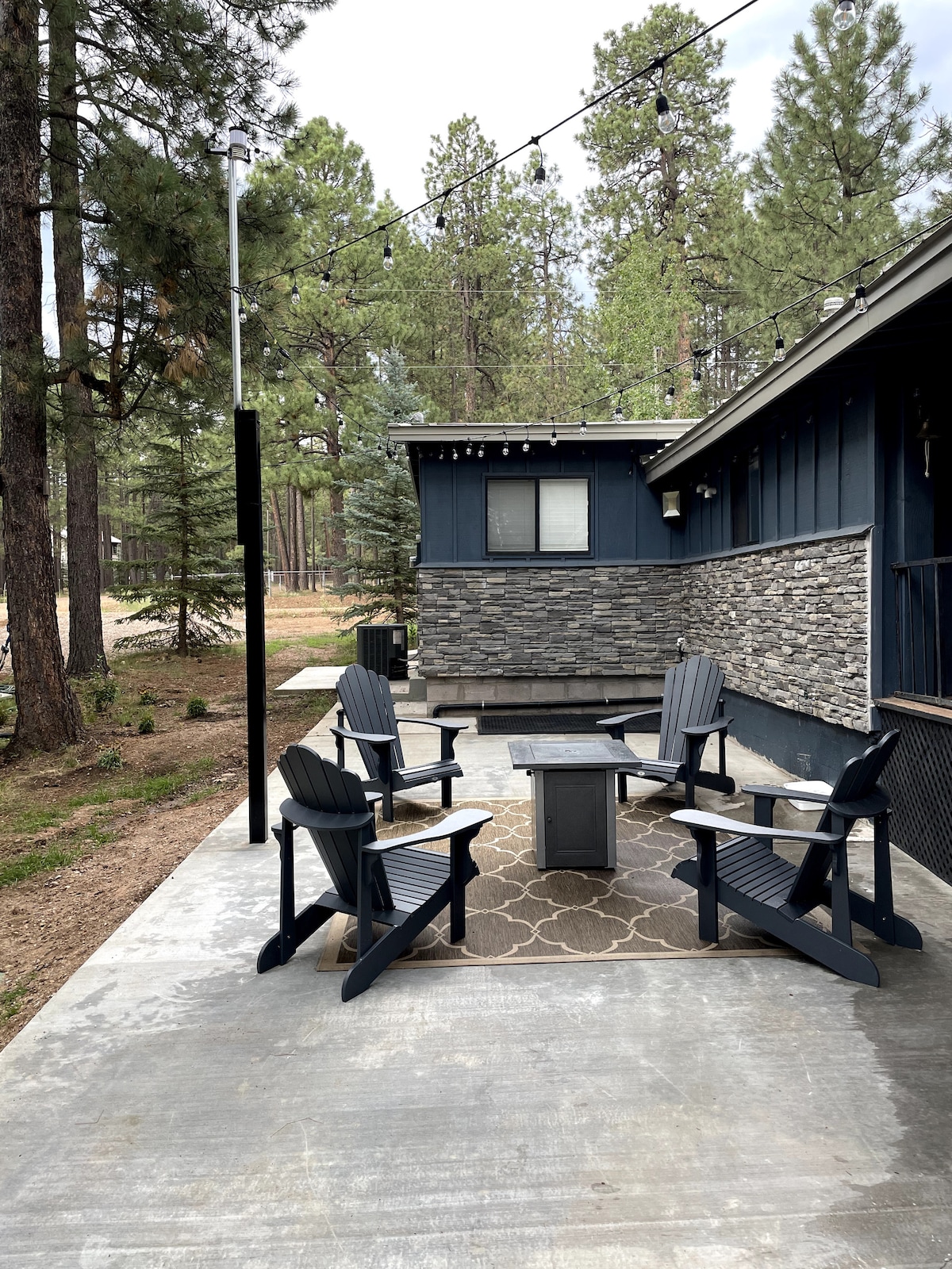 An outdoor seating area is set with four dark wood adirondack chairs surrounding a small table on a concrete patio. A patterned rug lies beneath the table, and trees are visible in the background, creating a serene atmosphere.