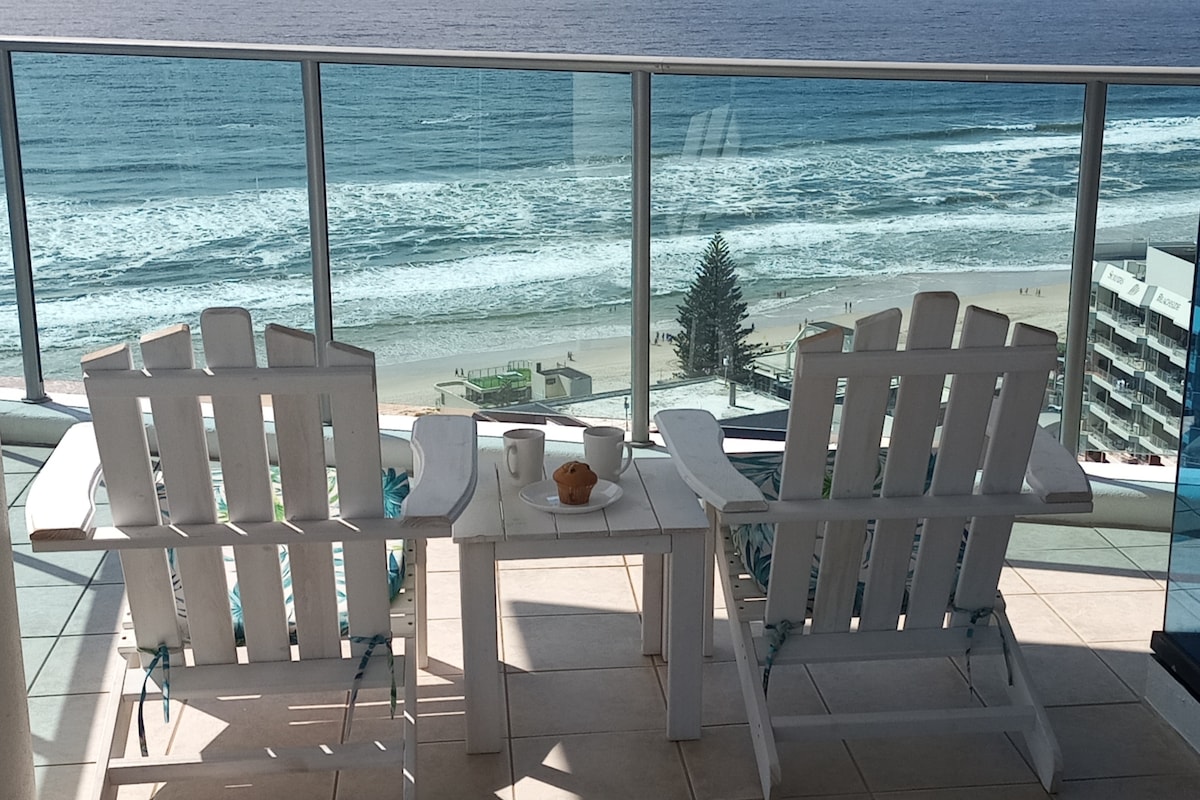 Two white deck chairs face the ocean, positioned on the balcony. A small table between the chairs holds a cup and a pastry. The vast beach and rolling waves are visible beyond the glass railing.