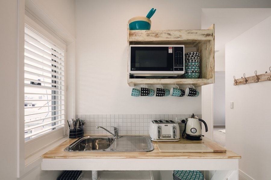 A compact kitchen is equipped with essential appliances, including a microwave, kettle, and toaster, all positioned on a wooden shelf. A stainless steel sink and a neatly arranged set of utensils contribute to a functional culinary space. Natural light streams through a nearby window with white blinds.