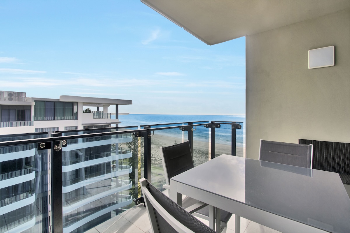 A balcony view showcases a clear blue sky above the ocean, with gentle waves lapping against the shore. A modern outdoor dining table is set with four chairs, providing a perfect space for enjoying meals and the scenic coastline.