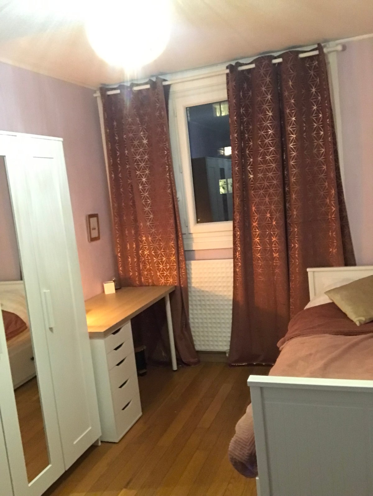 A spacious bedroom contains a single bed that can convert into a double. A desk is positioned beside a wardrobe, with warm lighting filtered through the sheer curtains. Wooden flooring complements the soft color palette of the room.