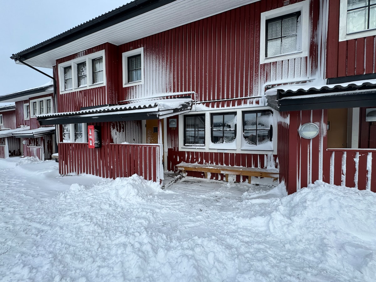 The exterior of a red wooden building is visible, adorned with white snow covering the ground and roof. Large windows allow natural light to enter, and a bench is positioned outside the entrance. The environment reflects a winter landscape.