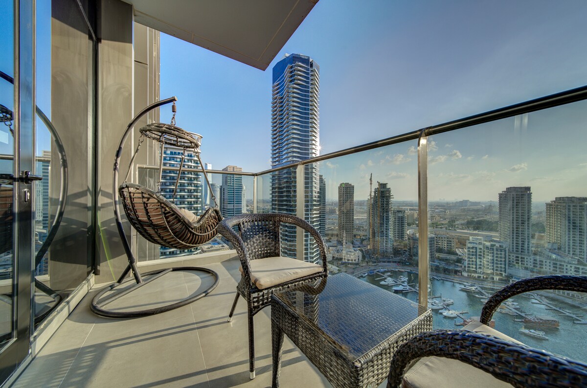 A balcony features a suspended hanging chair and a small wicker table with two chairs, providing a comfortable space for relaxation. The expansive view includes the marina and surrounding skyscrapers under a clear blue sky.