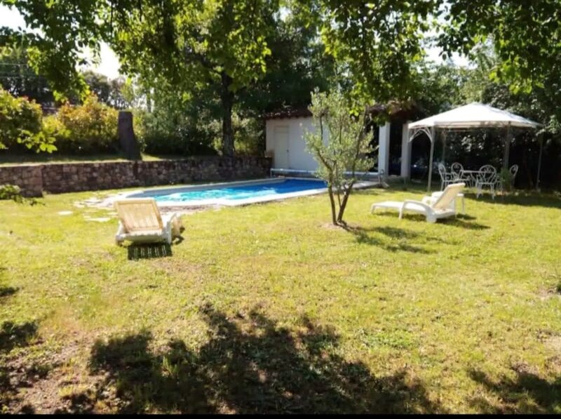 A serene outdoor area is highlighted by a well-maintained pool surrounded by grass and trees. Comfortable lounge chairs are positioned nearby, offering a spot to relax. A shaded seating area with a table and chairs is visible under a canopy, creating a peaceful setting.