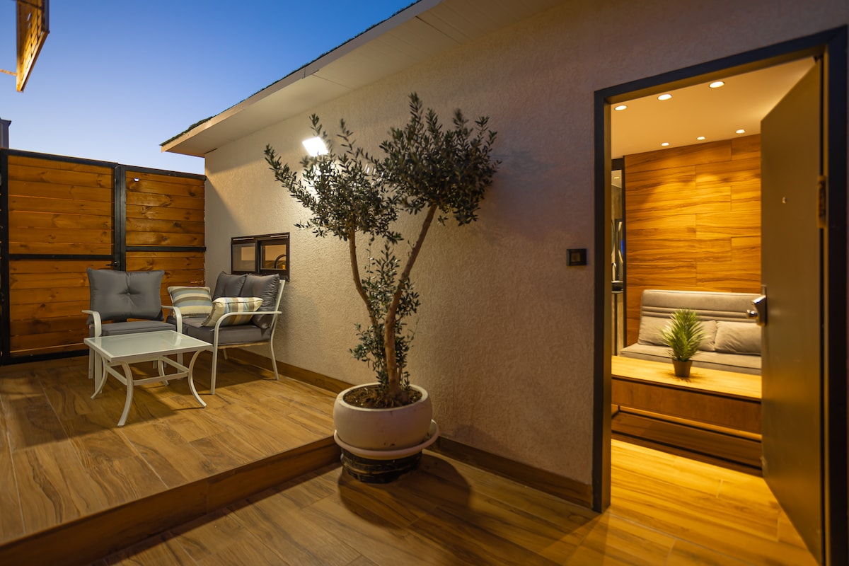 An inviting outdoor space features a comfortable seating area with two chairs and a side table, accompanied by a potted plant. Soft lighting illuminates the space, while a door leads to the interior, showcasing a warm wooden accent wall.