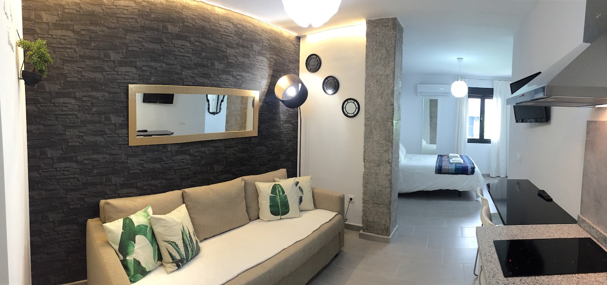 A modern living area features a light-colored sofa adorned with leafy throw pillows. A large mirror reflects the space, while dark stone accents contrast with the bright walls. In the background, a spacious bed is visible, accompanied by soft lighting and air conditioning.