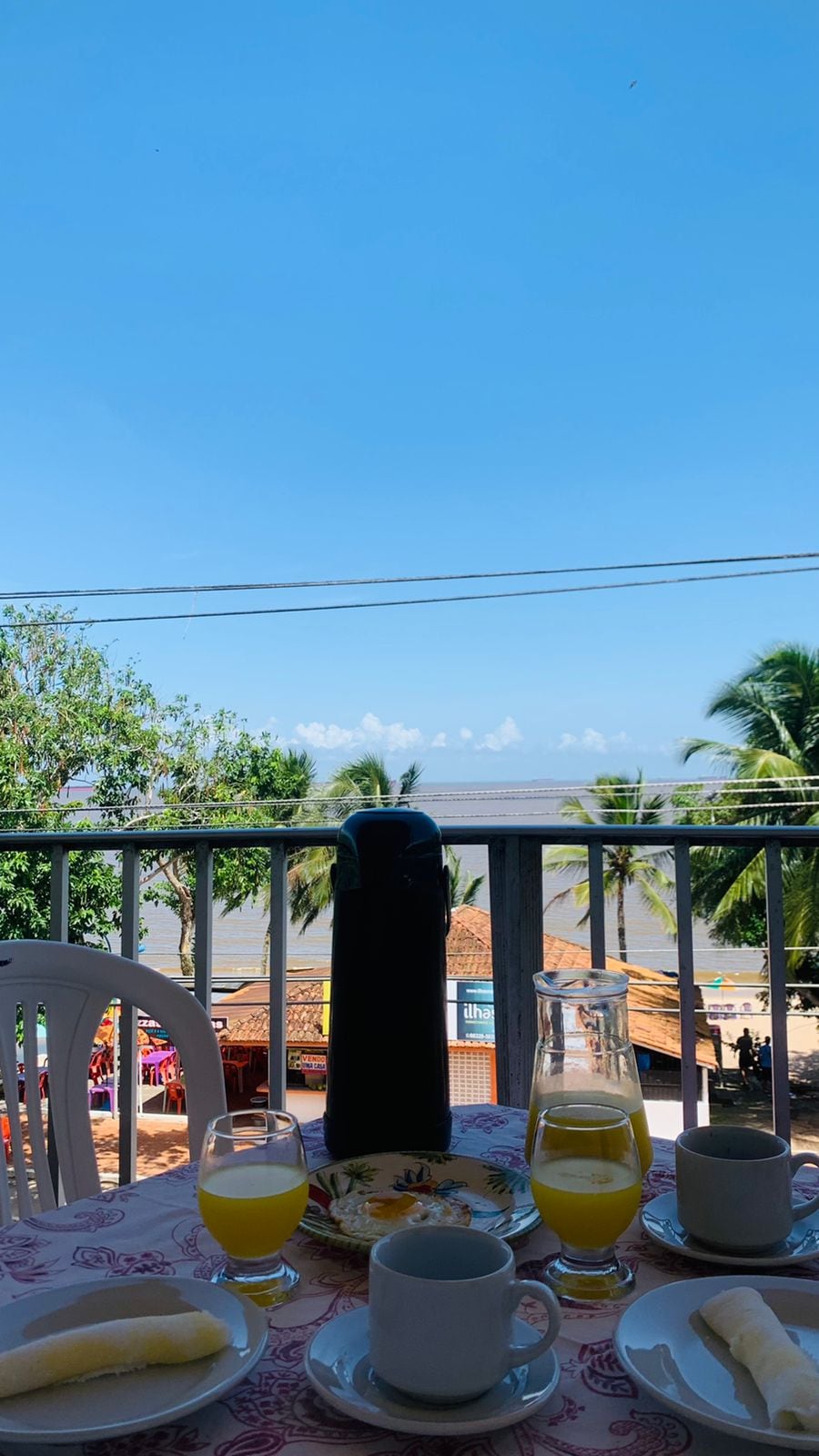 A serene balcony view is captured, featuring lush palm trees and a distant beach. A table is set with glasses of yellow drink and a thermos, alongside a small plate of bananas. The bright blue sky adds a cheerful tone to the scene.