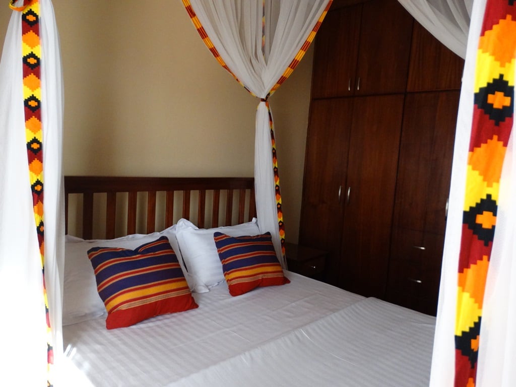 A spacious bed is framed by a decorative mosquito net with vibrant patterns. Two pillows with bold stripes are positioned at the head of the bed. A wooden wardrobe stands in the background, providing ample storage space.