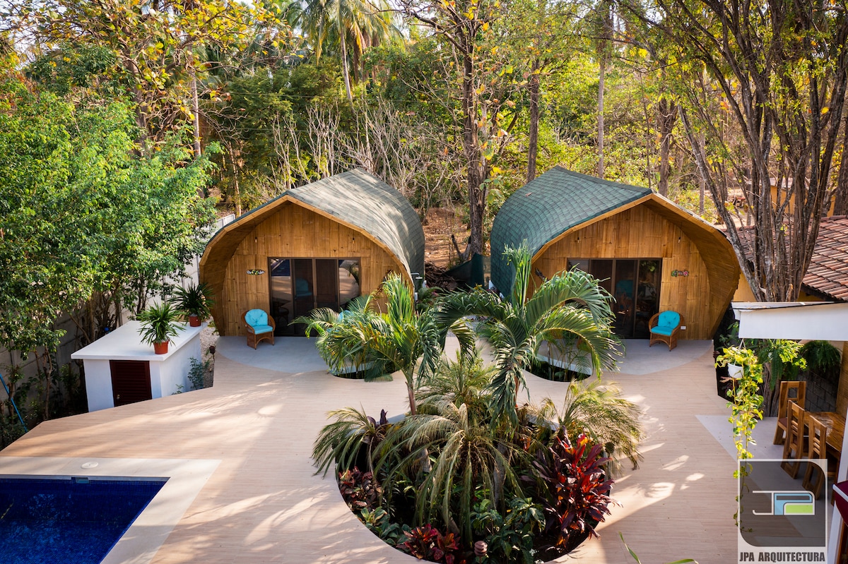Two wooden chalets with distinctive curved roofs are surrounded by lush greenery. Each chalet features large glass doors that provide views of the nature outside. A pool is visible in the foreground, bordered by a wooden deck and vibrant tropical plants.