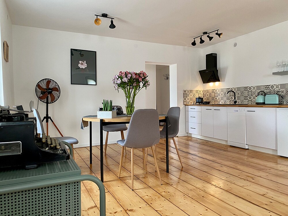 A modern kitchen area is displayed, featuring light wood flooring and white cabinetry. A round dining table is surrounded by four gray chairs, while a bouquet of flowers adds a touch of color. Vintage decor is also visible, including an old-fashioned fan and typewriter.