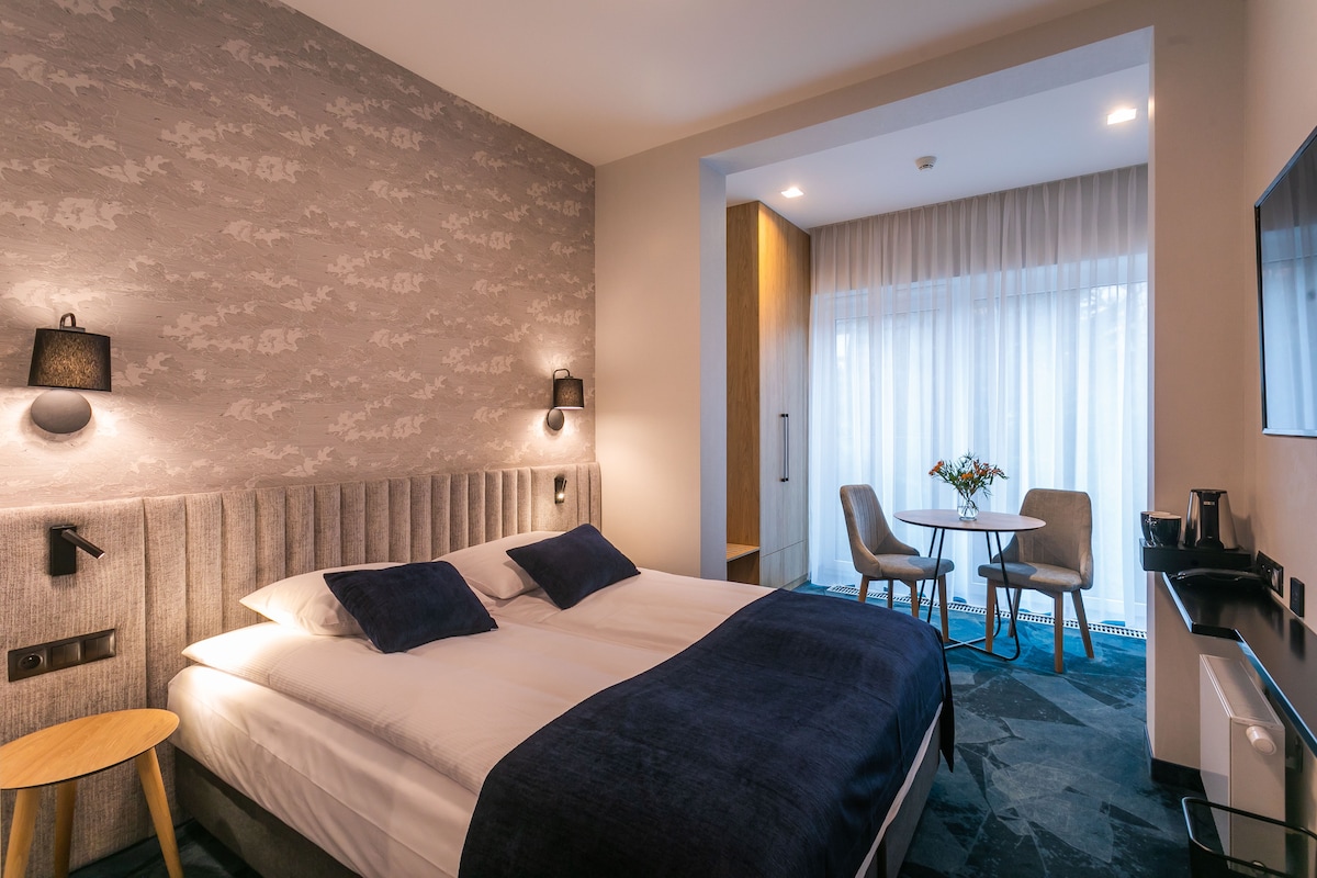 A neatly arranged room features a spacious queen bed with soft bedding, against a textured wall. A small dining area with two chairs and a table is located near the window, where natural light filters through sheer curtains.