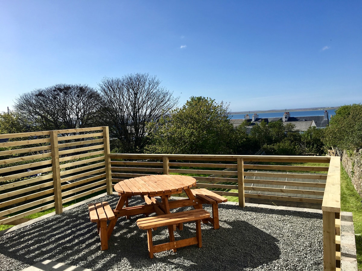 An outdoor seating area is visible, featuring a round wooden table surrounded by benches. The space is set on a gravel surface, framed by wooden slatted fencing. In the background, trees and the distant ocean can be seen under a clear blue sky.