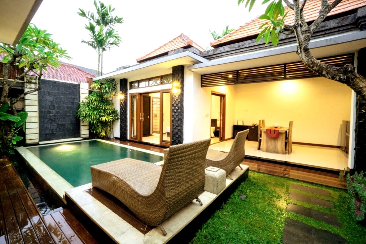 A private pool is surrounded by a garden, with lounge chairs positioned nearby. The contemporary villa features large glass doors that provide views of the open-plan living area, where a dining table is set. Lush greenery complements the modern design.