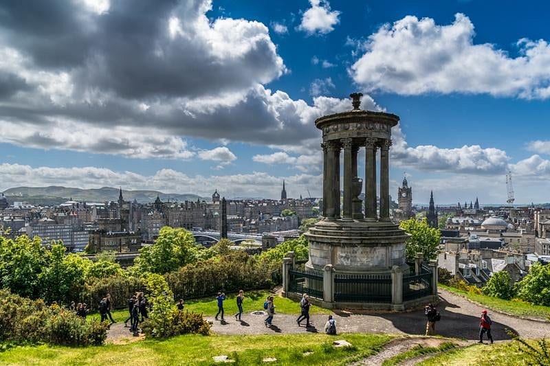 An expansive view of Edinburgh is showcased, featuring a prominent monument at the forefront surrounded by lush greenery. A group of people is seen walking along a path, with a backdrop of the city’s distinctive skyline, detailed with towers and historic buildings under a partly cloudy sky.