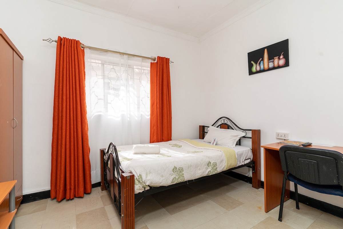 A neatly arranged private room features an individual bed with a light-colored blanket and towels neatly folded on top. A window partially draped with sheer curtains allows natural light to enter. An orange curtain adds a pop of color beside the bed, while a desk and chair occupy one corner.