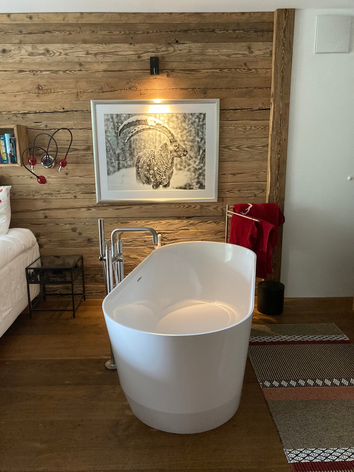 A freestanding white bathtub is positioned against a textured wooden wall, complemented by a framed black and white photograph featuring a snow-covered scene. Nearby, a red towel hangs on a rack, enhancing the bathroom's feature while a stylish rug lies beneath.