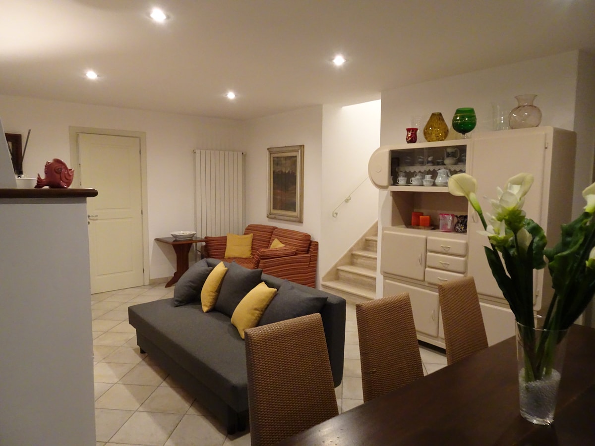 A spacious living area is presented, featuring a gray sofa adorned with yellow cushions. A dining table with woven chairs is visible in the foreground, while shelves displaying decorative items are arranged against the wall. Soft lighting enhances the comfortable atmosphere, complemented by a staircase leading to an upper level.