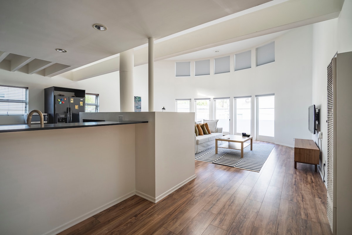 An open-concept living area is revealed, featuring large windows that allow natural light to illuminate the space. A cozy seating arrangement is positioned around a minimalist coffee table, with a television mounted on the wall opposite. Light wood flooring contrasts with the white walls.