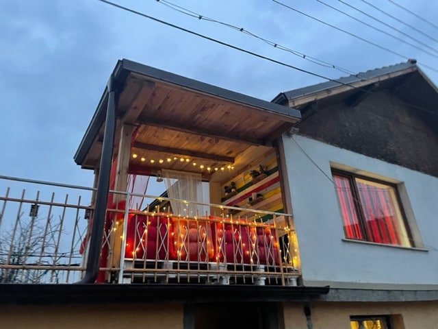An outdoor balcony is highlighted with string lights, creating a warm ambiance. The balcony features seating with vibrant cushions and is adorned with potted plants. The surrounding structure displays a mix of wooden and painted surfaces, with large windows offering a view of the interior.
