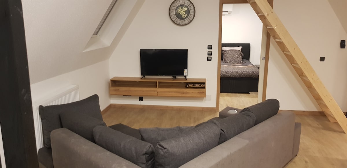 A living area with a gray sectional sofa is positioned near a wooden media unit, which supports a flat-screen television. A clock is mounted on the wall, and a staircase leading to the upper level is visible. In the background, a cozy bedroom is glimpsed.