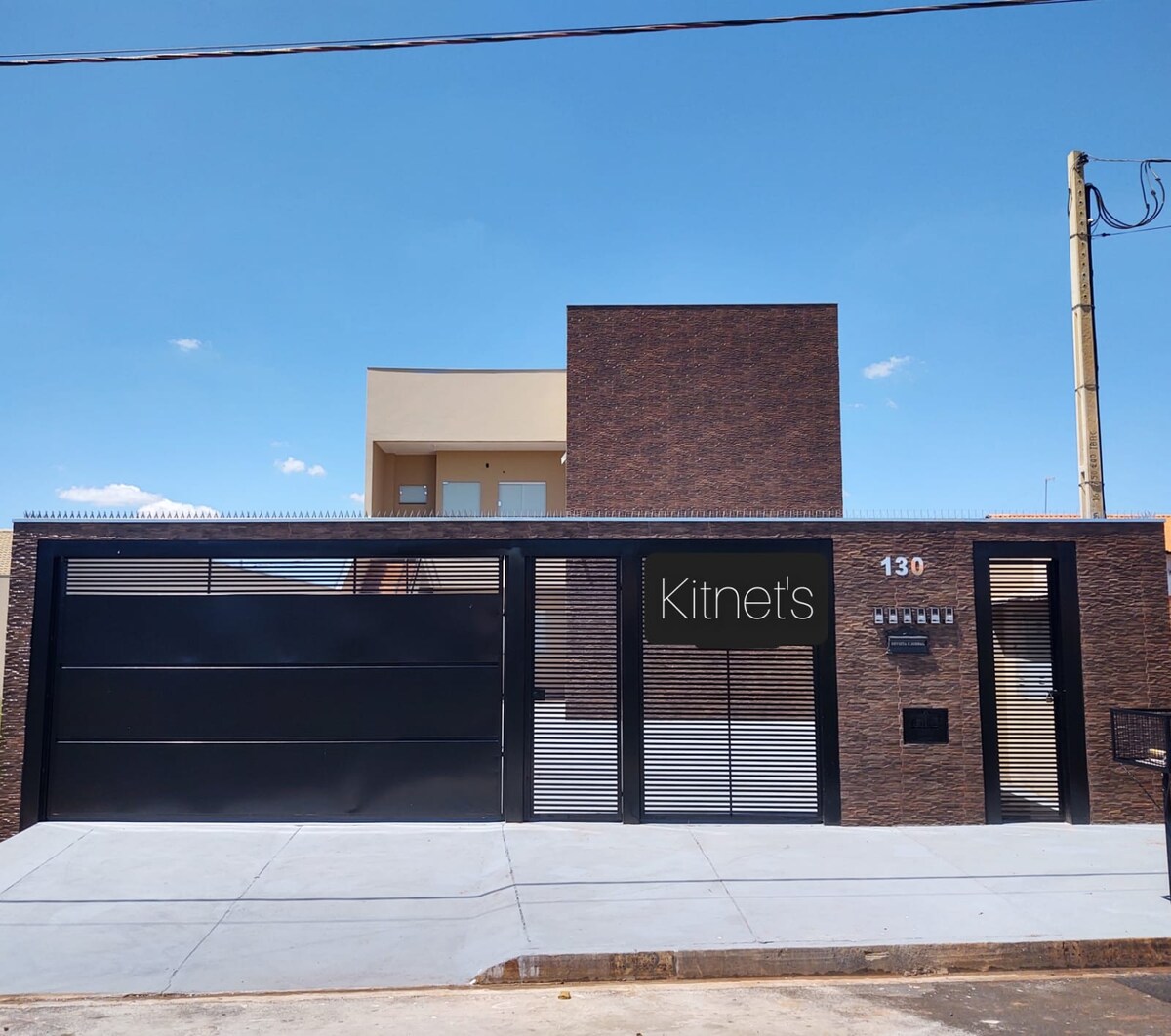 The exterior of the building showcases a modern, minimalist design with a sleek black gate and a contrasting brick facade. Large windows provide views into the interior spaces, while the clear blue sky creates a bright backdrop.