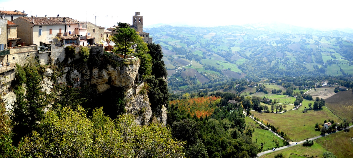 A panoramic view captures the medieval village of Montefalcone perched on a rocky cliff, surrounded by lush green hills and valleys. The landscape features a mix of fields and wooded areas, under a gentle sky. The stone buildings of the village blend seamlessly with the natural surroundings.