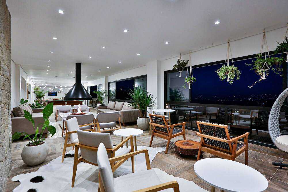 An open lounge area features modern seating arrangements, including light wood chairs and small white tables. Large windows frame the view of the outside, while hanging planters add a touch of nature. A stylish black fireplace serves as a focal point in the space.