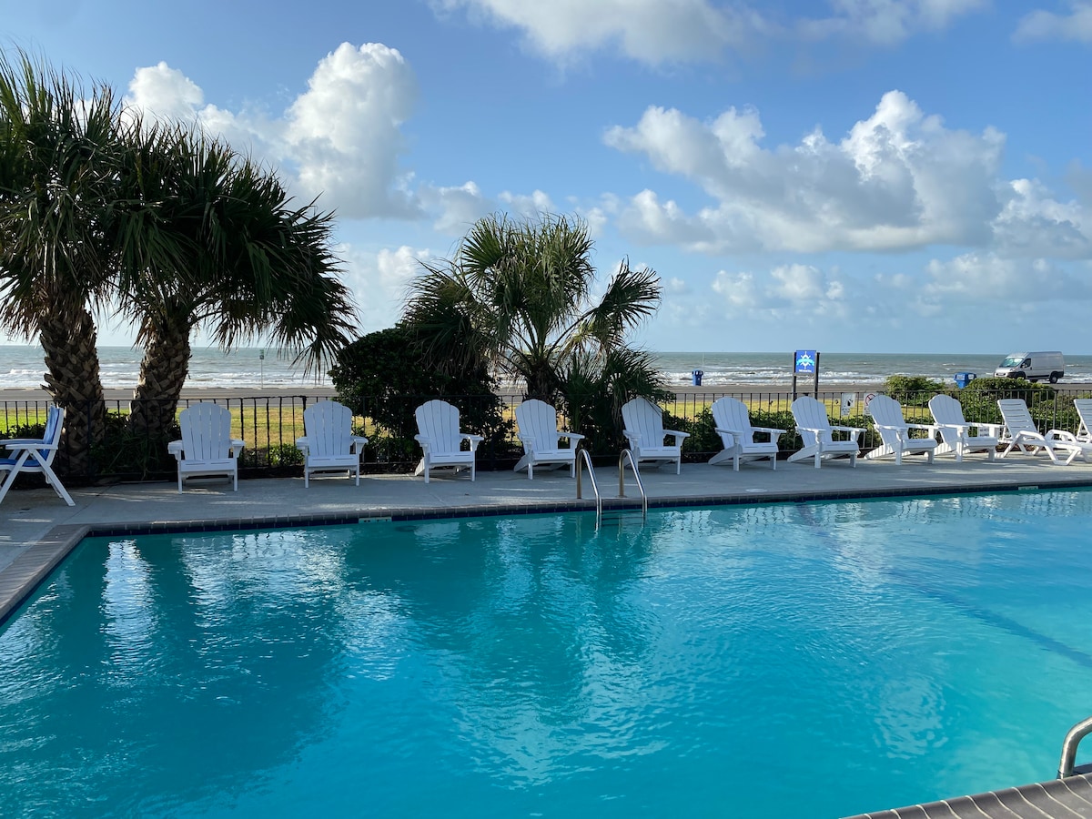 A clear blue swimming pool is framed by palm trees and lounge chairs. The ocean can be seen in the background, reflecting the sky's soft clouds. The inviting pool area provides a pleasant space for relaxation.