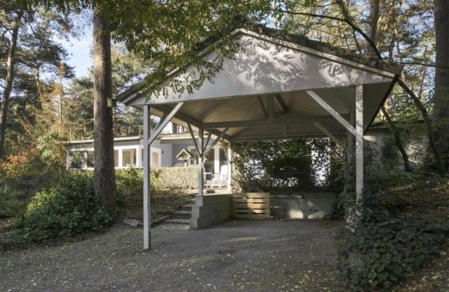 A covered carport provides shelter in front of the wooden house, surrounded by lush greenery and tall trees. Steps lead from the carport to the entrance of the house, where large windows invite natural light into the living space.