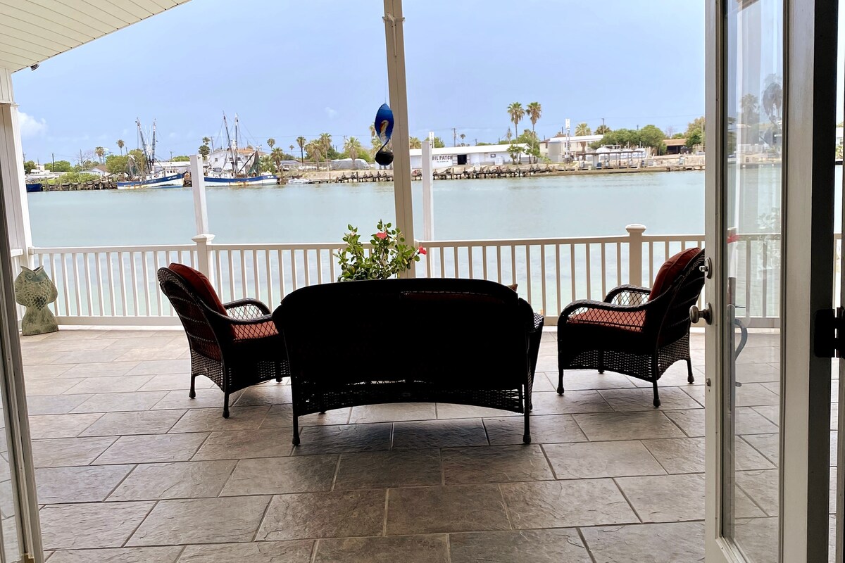 A covered patio area presents two wicker chairs and a curved love seat, facing a view of the Intracoastal Waterway. Decorative plants are positioned on the railing, enhancing the outdoor space. The calm water reflects a cloudy sky, framing the scene with passing boats in the background.