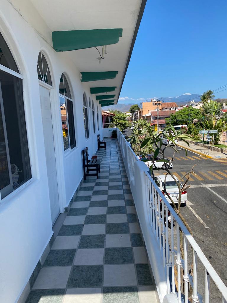 A wide corridor is lined with a checkerboard tile pattern, featuring several black wooden benches. Arched windows allow natural light to fill the space, while potted plants are visible along the railing. The view includes nearby street scenes and distant mountains under a clear blue sky.