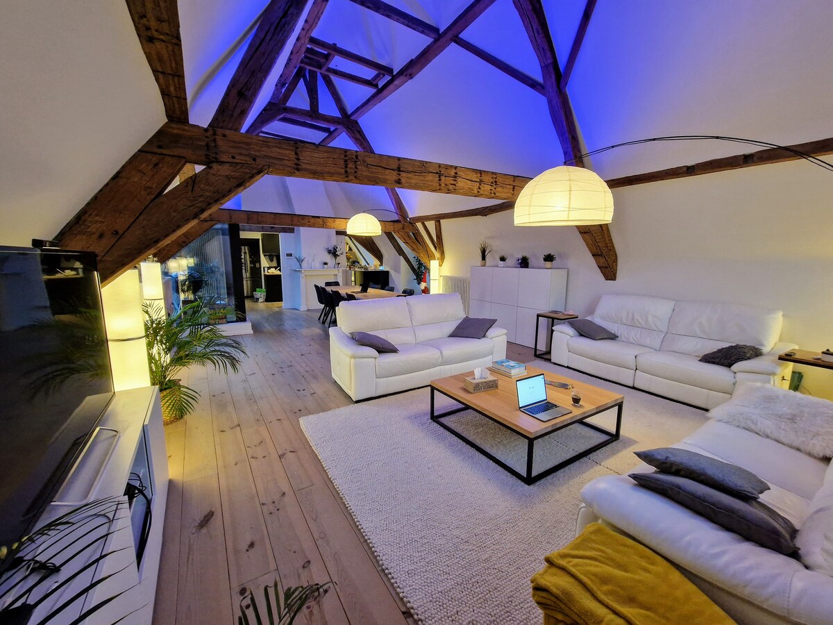 A spacious living room is displayed, featuring high vaulted ceilings with exposed wooden beams. Two large sofas in white surround a central coffee table, complemented by a soft rug. Soft, ambient lighting is provided by hanging lamps, enhancing the warm character of the room.