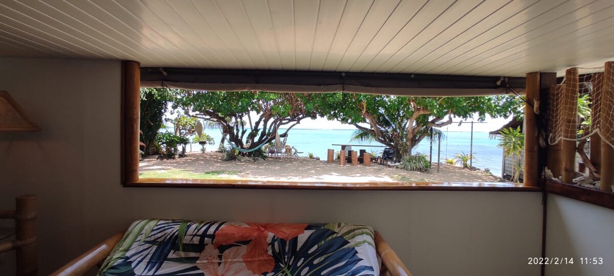 A large window offers a view of the beach and lagoon, framed by lush trees. The vibrant floral pattern of the bedding can be seen in the foreground, contrasting with the serene outdoor setting.