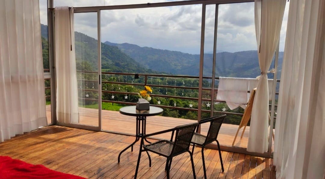 A spacious area is highlighted by large glass doors opening to a stunning mountain view. A wooden deck is furnished with a small table and two chairs. Sheer curtains frame the entrance, allowing natural light to fill the space.