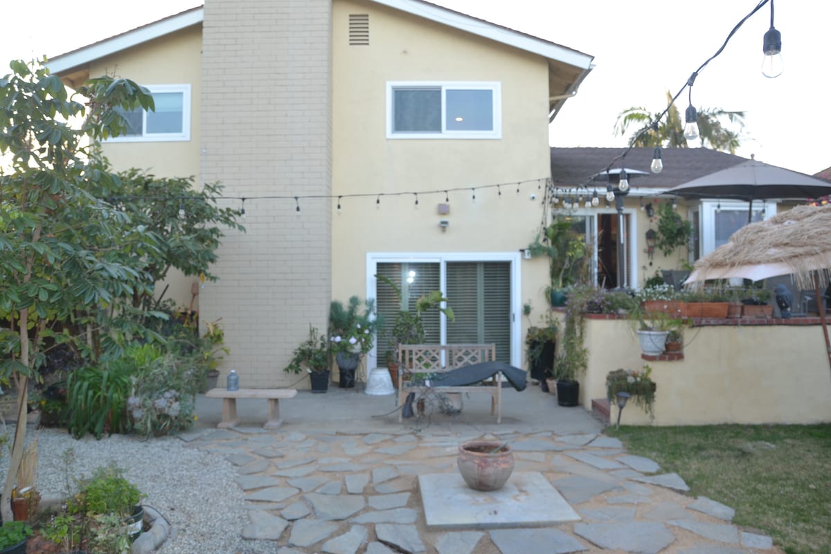The exterior of the home is showcased, featuring a two-story structure with large windows and a textured beige façade. A stone patio is visible, accompanied by a landscaped area with various plants. Decorative string lights hang overhead, adding a soft ambiance to the outdoor space.