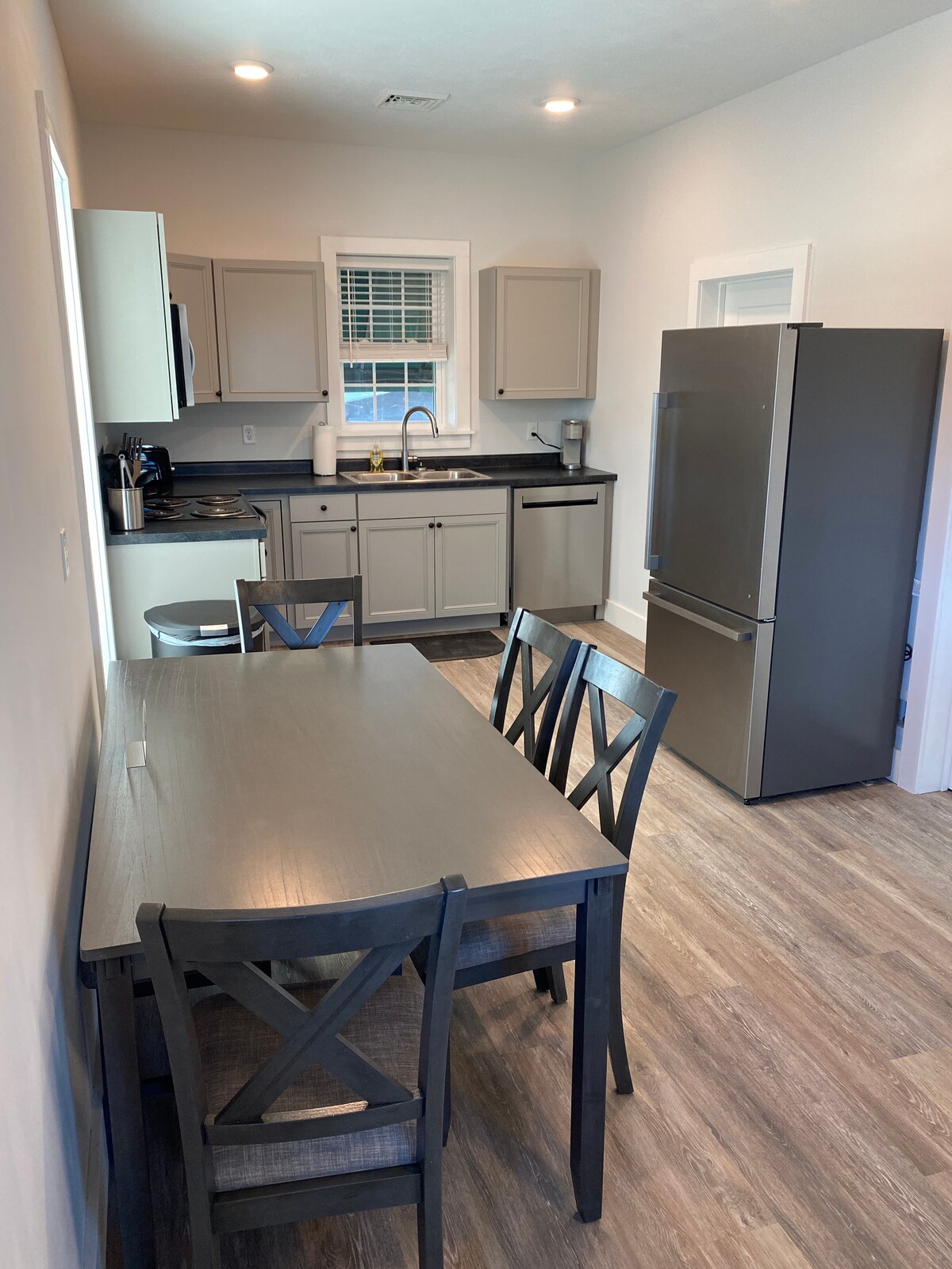 A bright kitchen area features modern cabinetry with a light grey finish, a refrigerator, and a stove. A dining table with four black chairs is positioned near a window, allowing natural light to filter in. The flooring provides a sleek wood-like appearance.