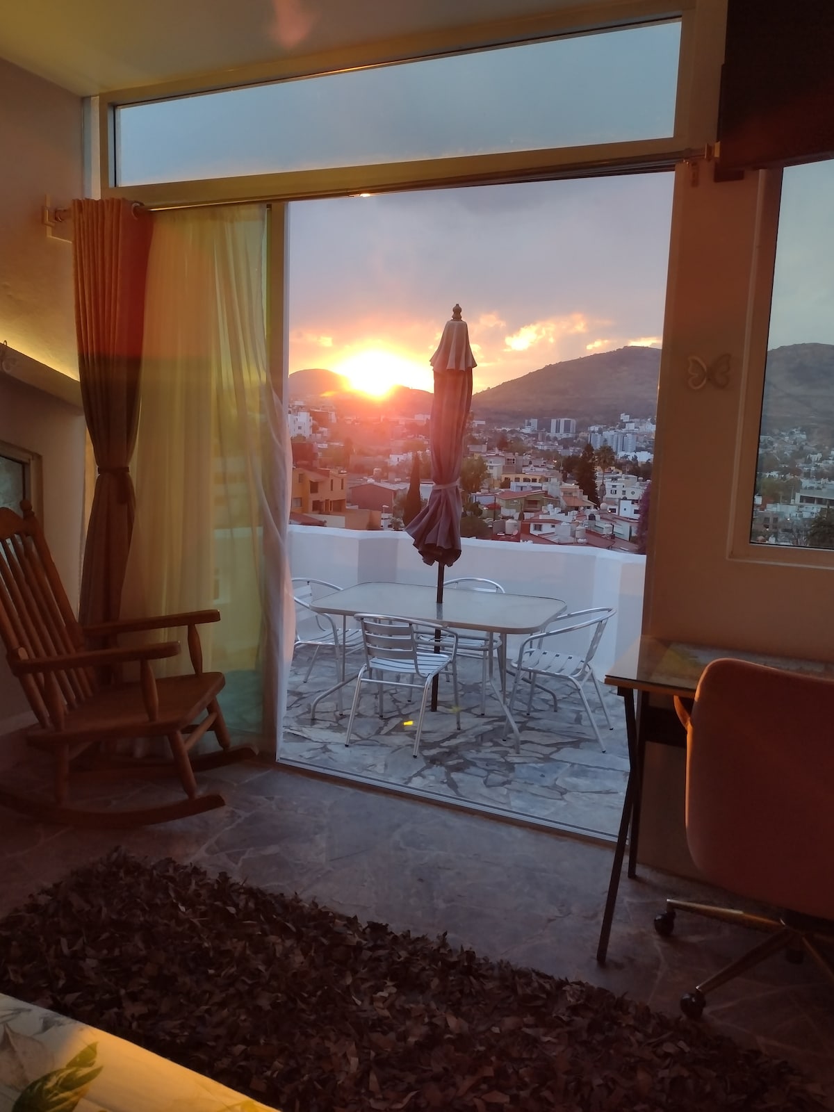 A spacious room features large glass doors opening to a private terrace with a table and chairs. A rocking chair is positioned indoors. The sunset casts warm hues, illuminating the distant mountains and the cityscape beyond.