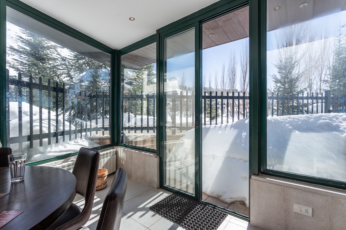 A sunlit sunroom features floor-to-ceiling glass panels, providing views of the surrounding snow-covered landscape. A wooden dining table is set nearby with dark chairs. A welcome mat lies at the entrance, and greenery from the outside complements the serene atmosphere.