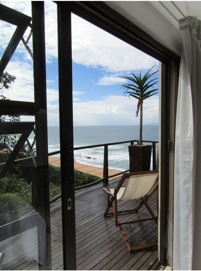 A private balcony is viewed through open glass doors, featuring a wooden deck and a single chair. A potted plant adds greenery, while the ocean stretches out beyond with gentle waves. A mix of sky and clouds is visible above the water.
