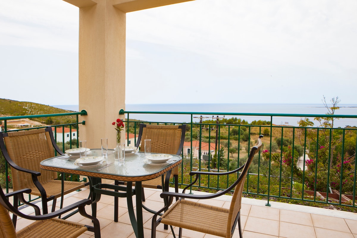 A balcony table set for four is seen, featuring a glass top and woven chairs. A scenic view of the sea and surrounding greenery is visible beyond the railing, with a hint of colorful foliage in the frame.