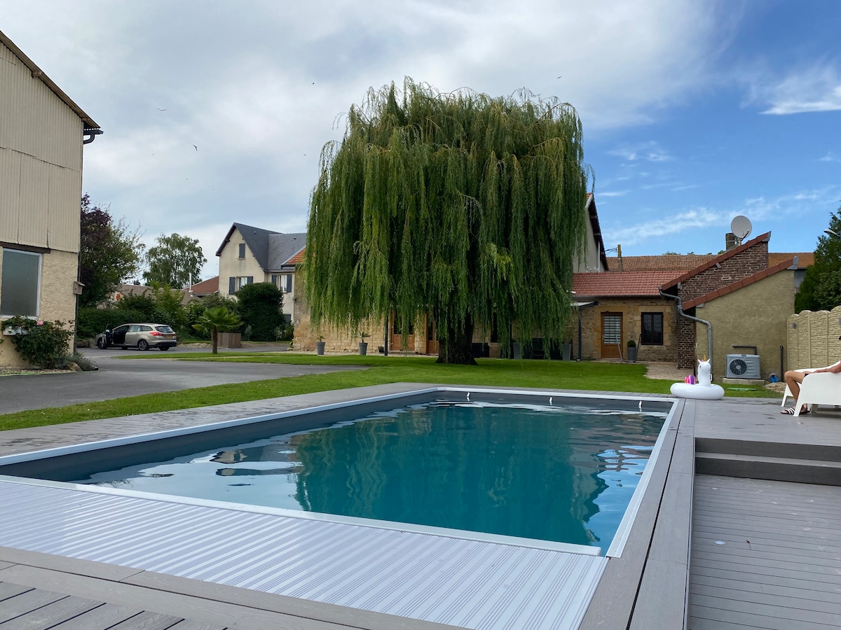 A swimming pool is set within a grassy area, bordered by a tall, leafy tree. Nearby buildings display a mix of architectural styles, including a low-rise structure with a traditional roof. The clear blue sky above complements the serene outdoor setting.