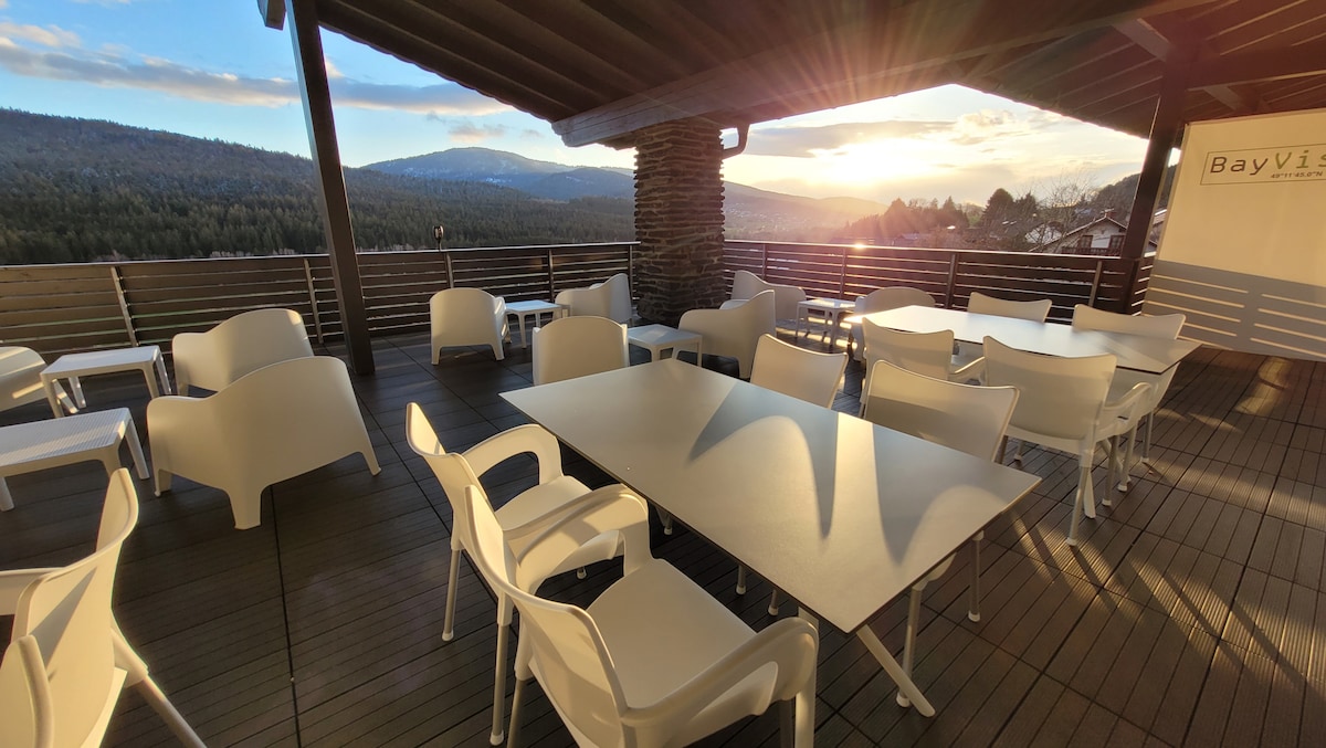 A spacious terrace is shown with several white chairs and tables arranged for dining. Large wooden beams support the overhead structure, and the surrounding mountains can be seen in the background, illuminated by a setting sun.