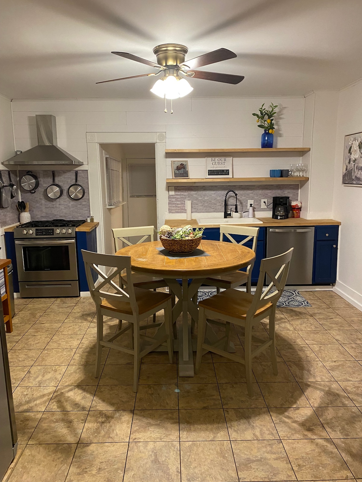 A bright kitchen area features a circular wooden table surrounded by four wooden chairs. A ceiling fan provides airflow, while stainless steel appliances are visible. Open shelving displays decorative items against blue cabinetry, complemented by a patterned rug on the tiled floor.