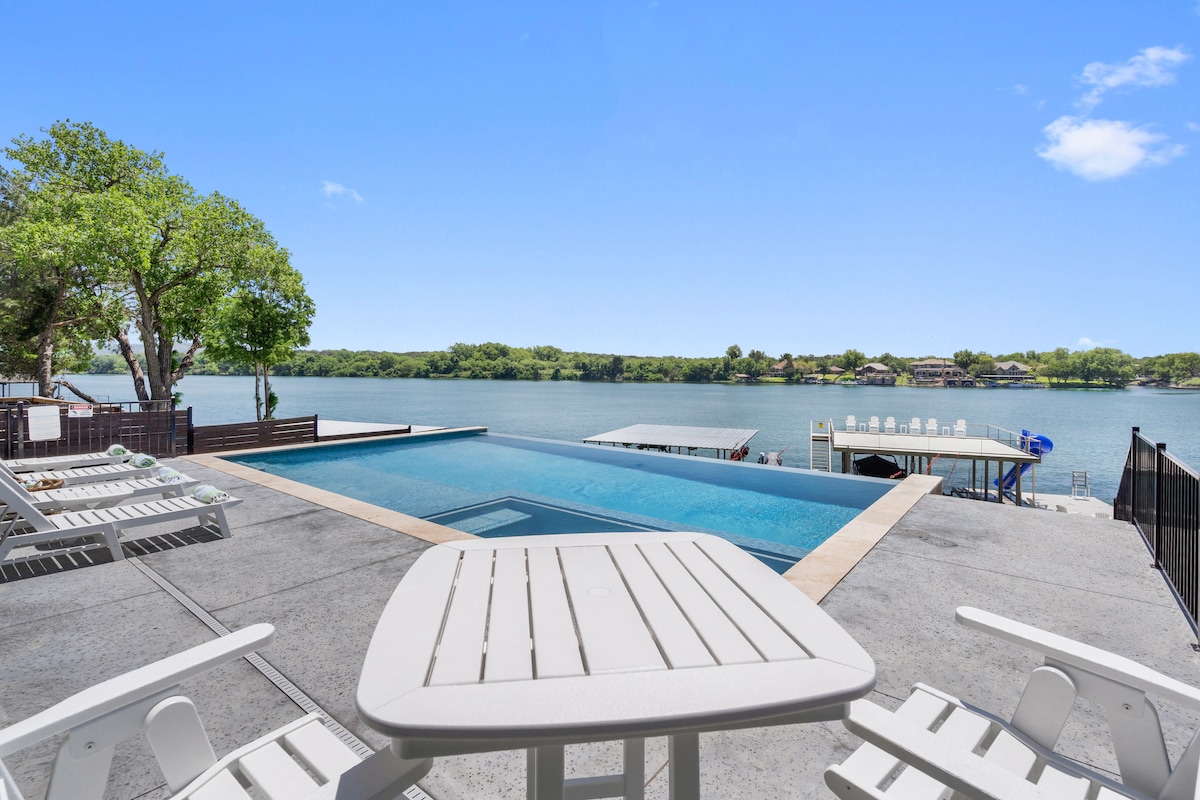 The outdoor area features an infinity pool overlooking Lake LBJ, surrounded by a spacious pool deck. Several lounge chairs are positioned for relaxation, and a private boat dock is visible, offering direct access to the water.