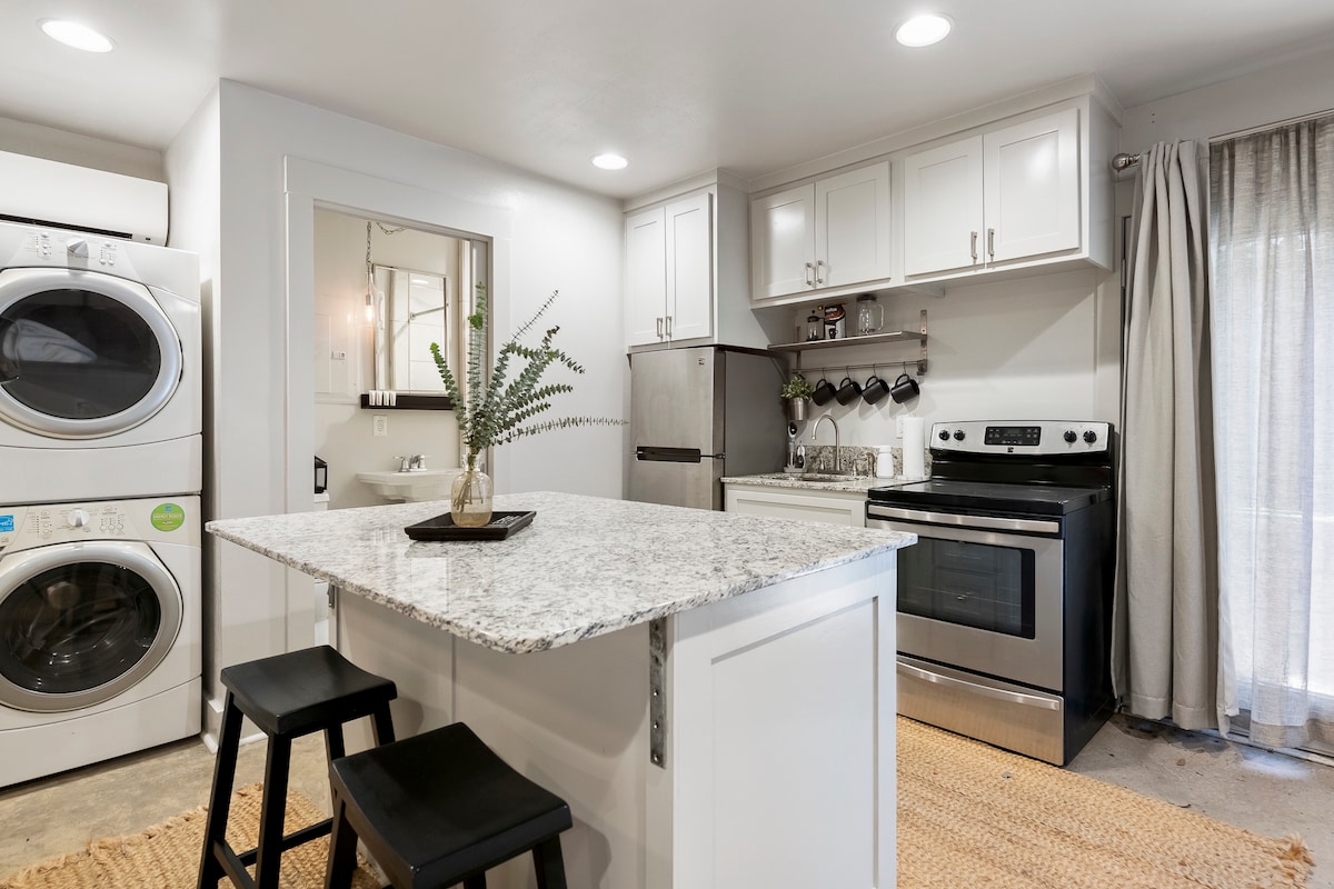 A modern kitchen is presented, featuring a central island with two stools. Stainless steel appliances, including a refrigerator and oven, are positioned along white cabinetry. A laundry area with stacked machines is visible. Natural light enters through a window adorned with light curtains.