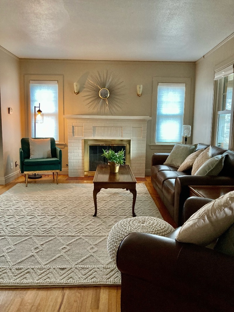 A cozy living room is presented with a brown leather sofa and a stylish armchair in green. A wooden coffee table sits on a textured area rug, while a simple decorative clock and wall sconces enhance the space. Natural light filters through two windows.